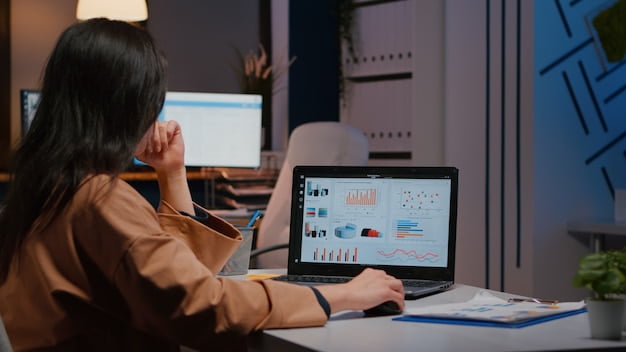 Overworked businesswoman typing financial statistics on laptop sitting at desk table in startup company office late at night